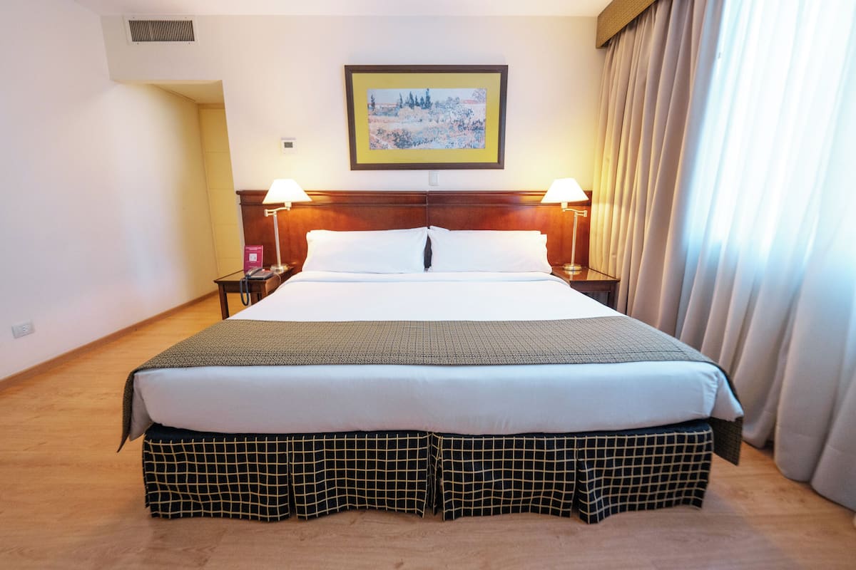 A hotel room with a large bed, two bedside lamps, a headboard, a framed picture on the wall, and a window with curtains.