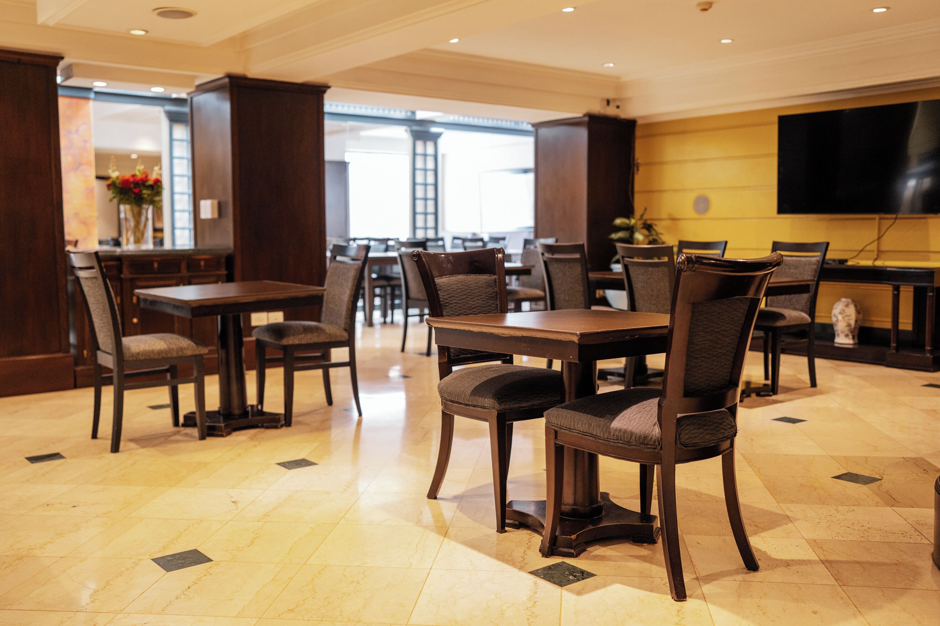 A hotel dining area with multiple tables and chairs, a large window, and a flat-screen TV mounted on the wall.