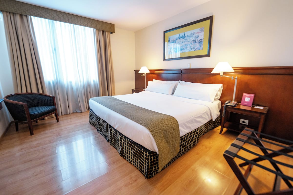 A hotel room with a large bed, two bedside tables, a chair, a window with curtains, and a framed picture on the wall.
