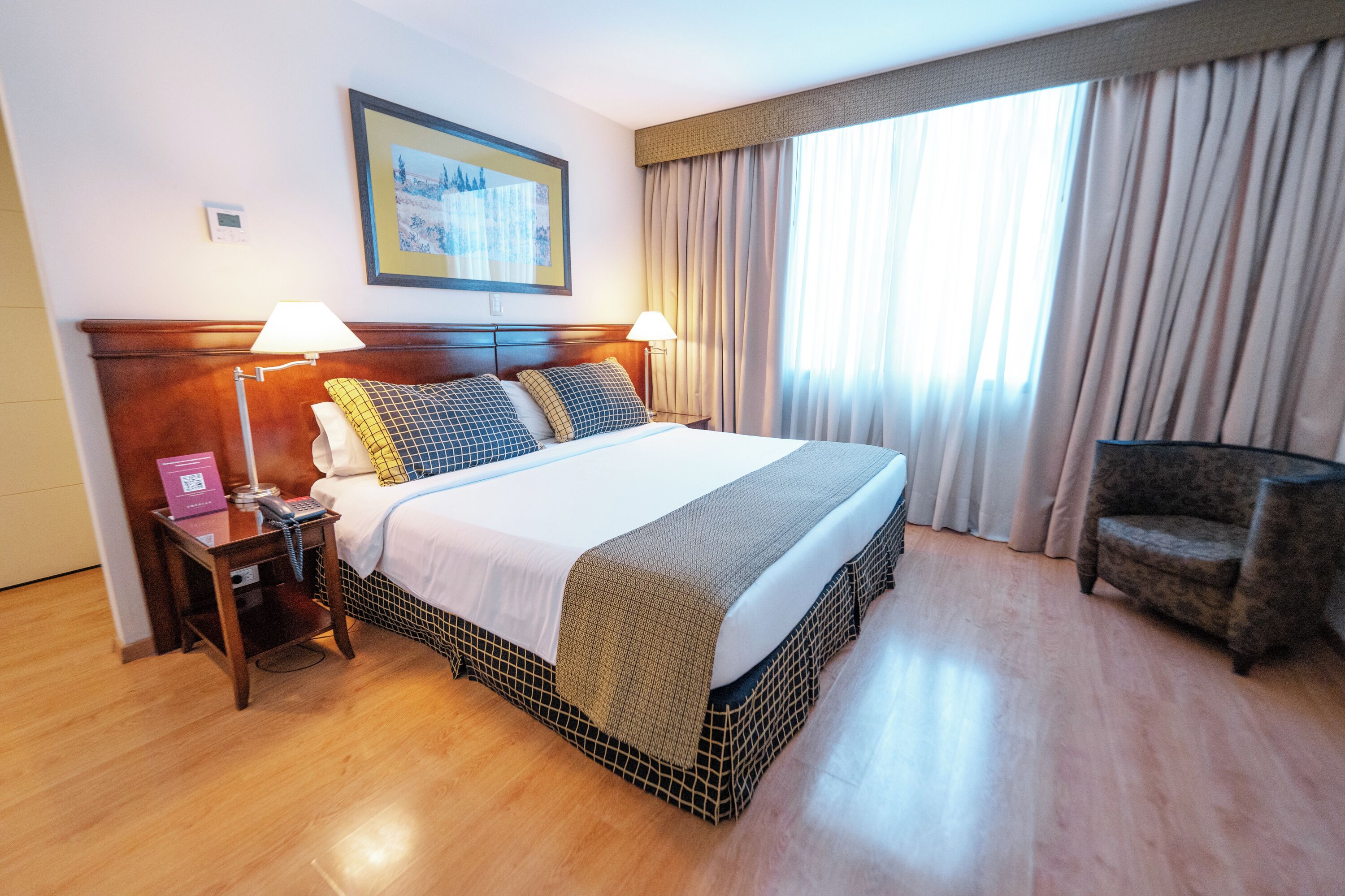 A hotel room with a large bed, two bedside lamps, a chair, a bedside table with a phone and a book, and a window with curtains.