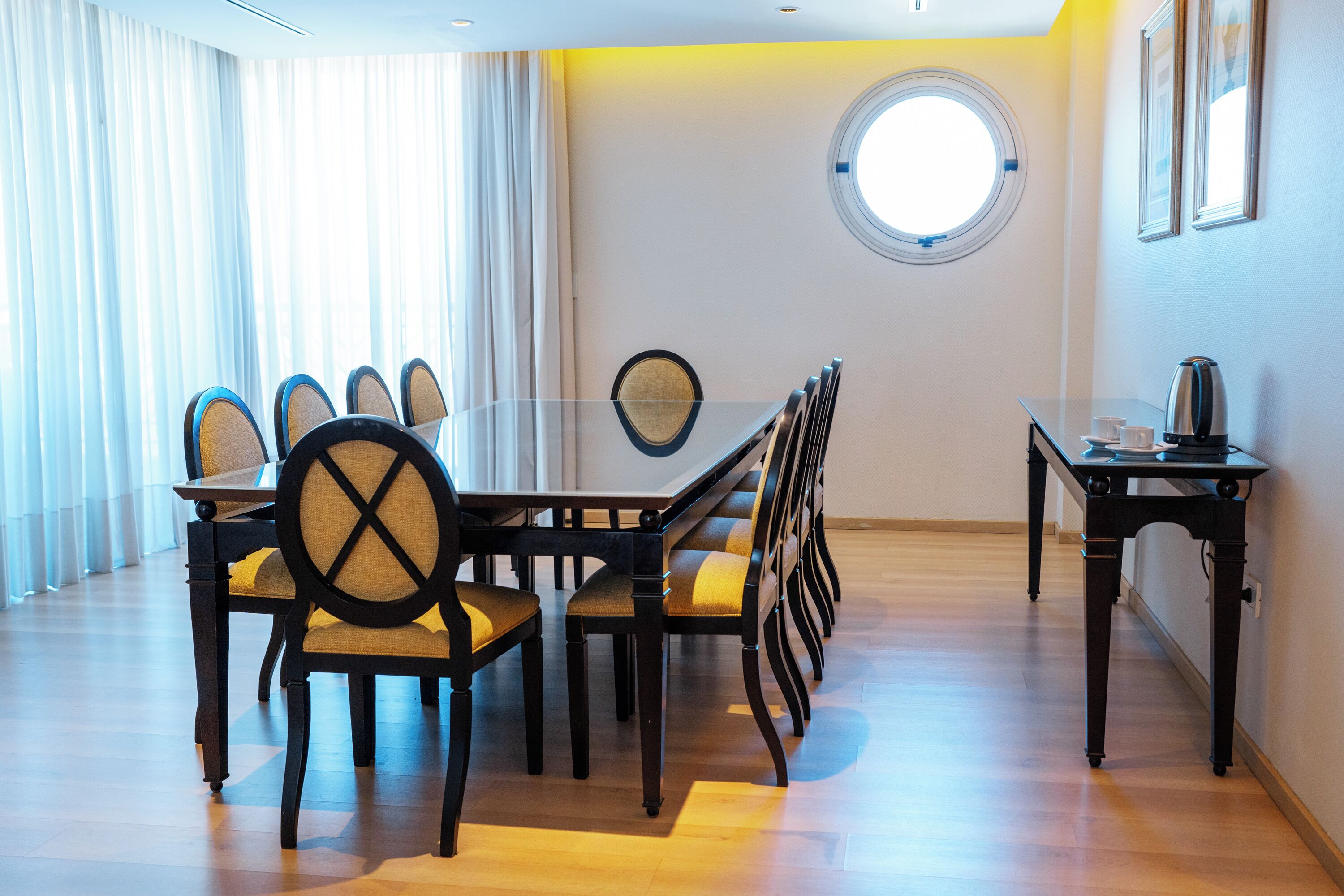 Een vergaderruimte met een glazen tafel, houten stoelen, een klein bijzettafeltje met een waterkoker en een raam met gordijnen.