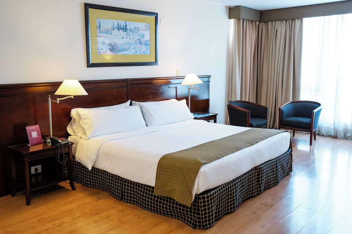 A hotel room with a large bed, two bedside lamps, a nightstand with a phone and a pink booklet, a chair, and a painting on the wall.