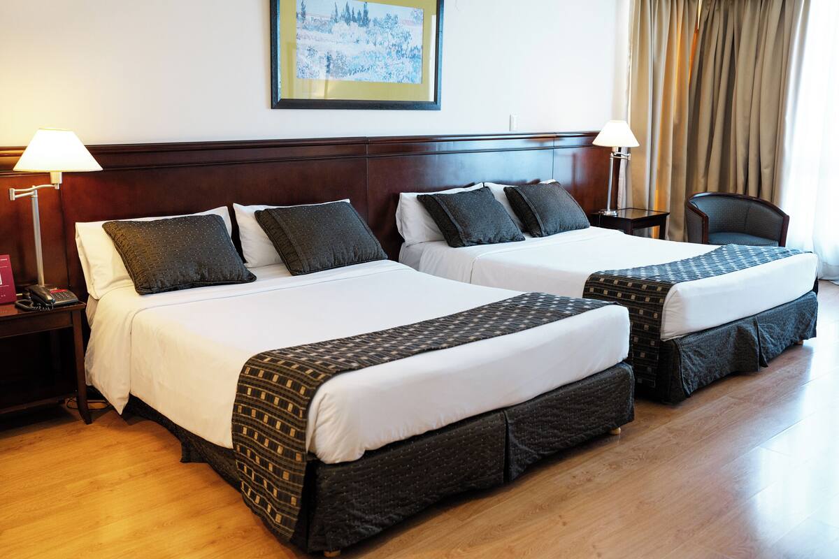 A hotel room with two beds, each with a patterned bedspread and pillows, a wooden headboard, a nightstand with a lamp, and a framed picture on the wall.