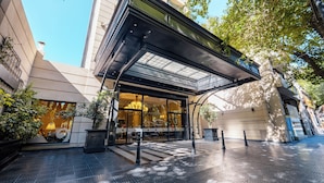 A hotel entrance with a glass canopy, a tiled pavement, and a reflection of a lamp in the glass.