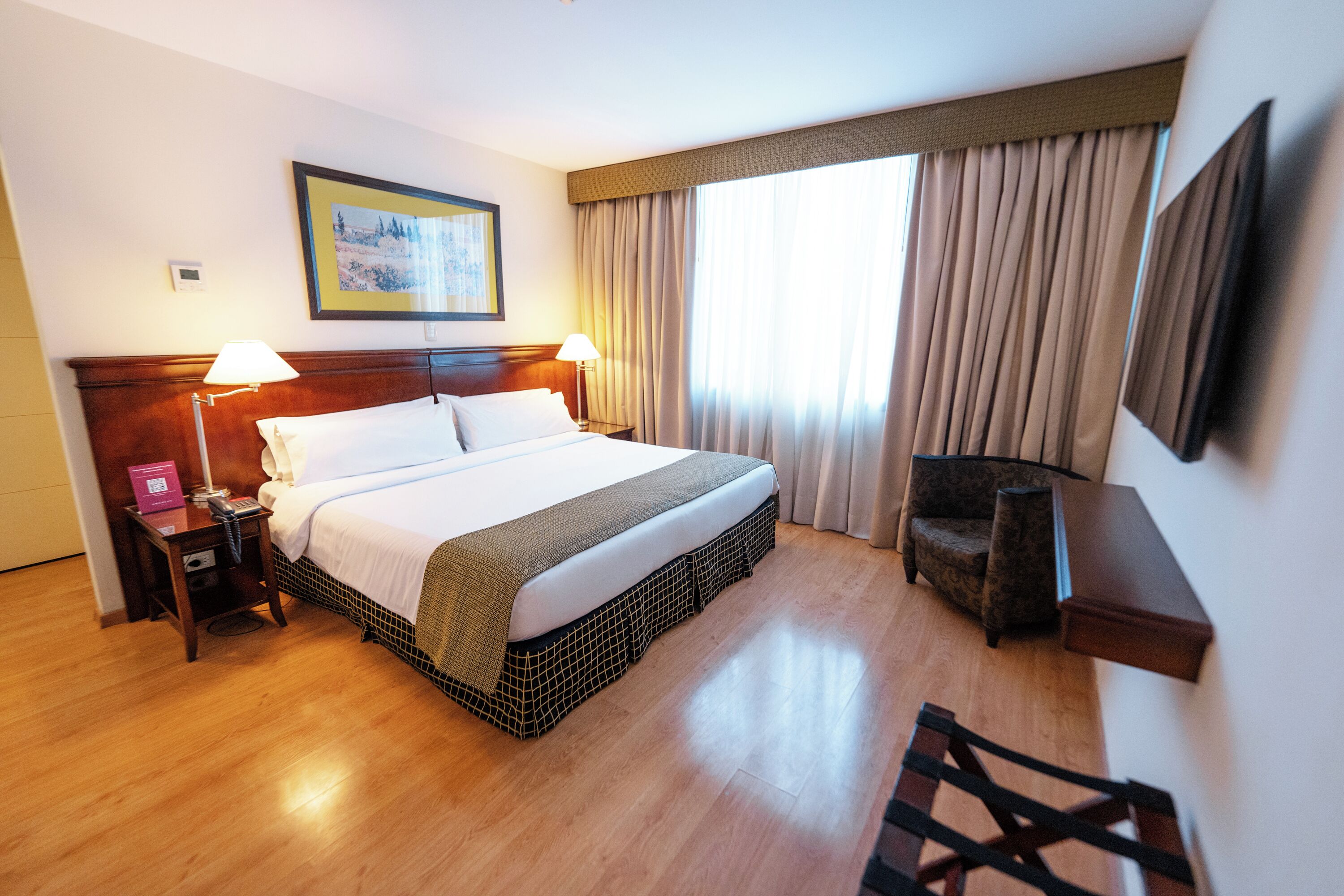 A hotel room with a bed, two bedside lamps, a chair, a TV, and a painting on the wall.