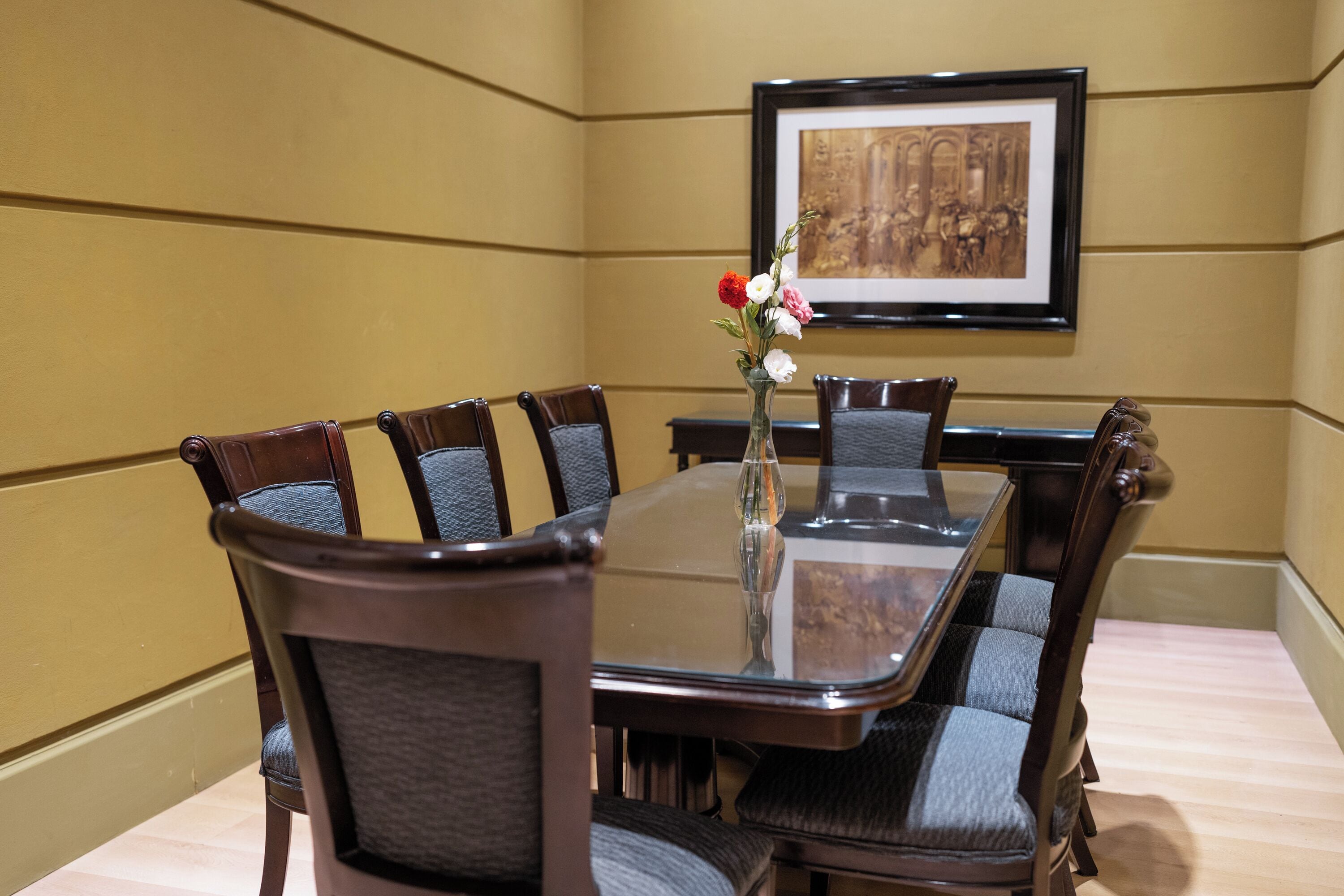 A conference room with a glass table, six upholstered chairs, a vase with flowers, and a framed picture on the wall.