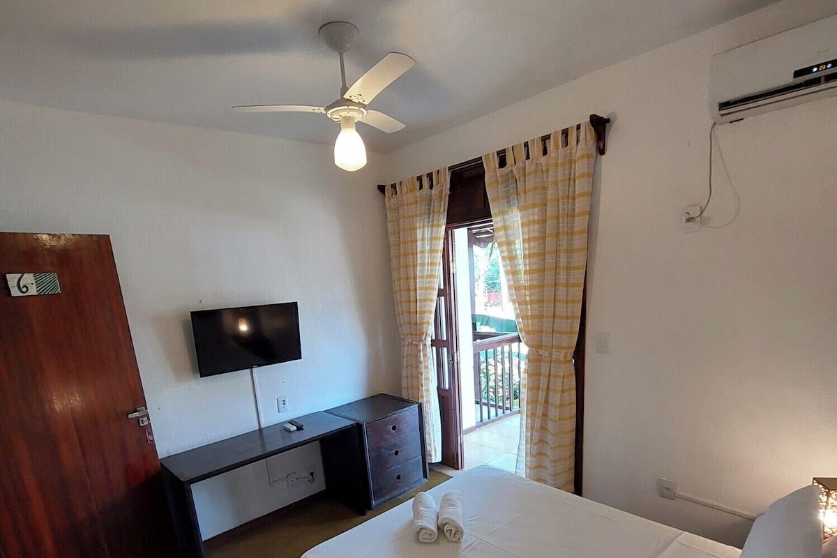 A bedroom with a bed, a desk, a television, a ceiling fan, and a window with curtains.