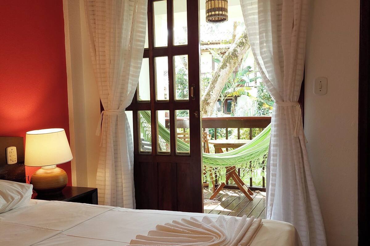A hotel room with a bed, a nightstand with a lamp, a window with curtains, and a view of a hammock and greenery outside.