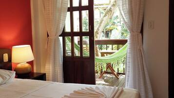 Quarto de hotel com cama, mesa de cabeceira com abajur, janela com cortinas e vista para uma rede e vegetação lá fora.