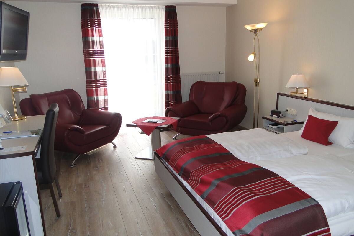 Une chambre d’hôtel avec un lit, deux fauteuils rouges, un bureau avec une lampe, une télévision et une table de chevet avec une lampe.