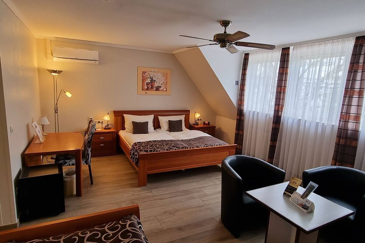 Une chambre d’hôtel équipée d’un lit, d’un bureau, d’une chaise, d’une petite table, d’un ventilateur de plafond et de rideaux.