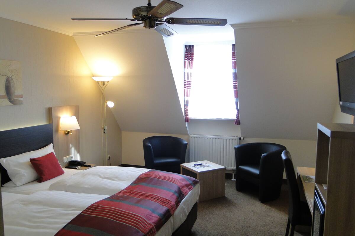 Une chambre d’hôtel avec un lit, deux chaises, une petite table, un ventilateur de plafond, une télévision et une fenêtre avec des rideaux.