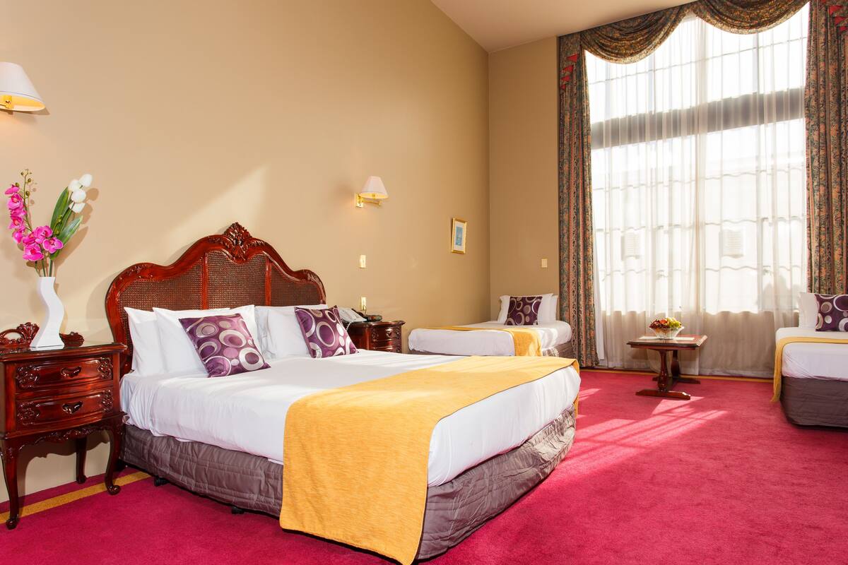 A hotel room with a large bed, a smaller bed, a nightstand, a lamp, a vase with flowers, and a window with curtains.