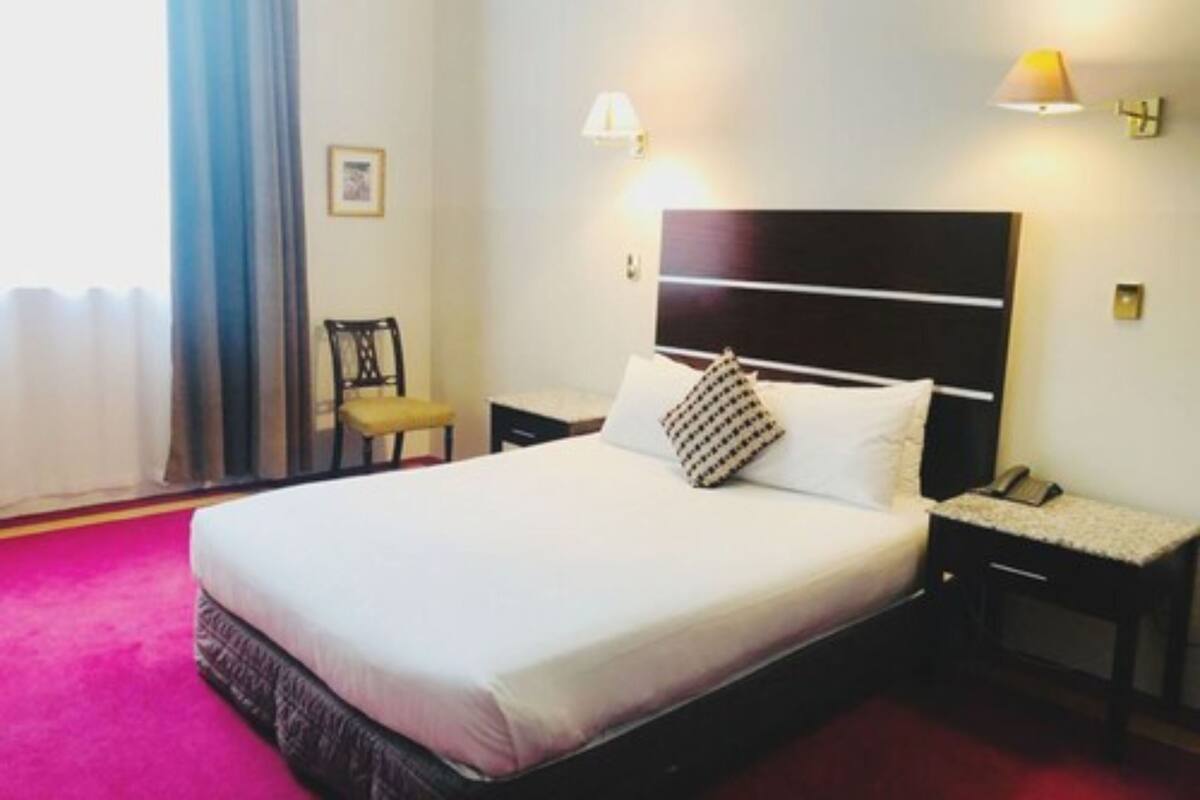 A hotel room with a large bed, a bedside table with a lamp, a chair, and a window with curtains.