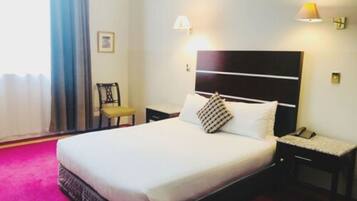 Een hotelkamer met een groot bed, een nachtkastje met een lamp, een stoel en een raam met gordijnen.