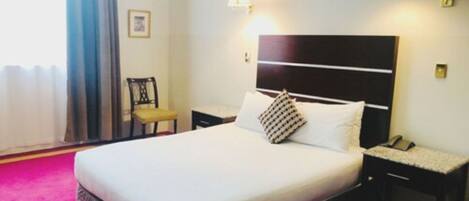 Una habitación de hotel con una cama grande, una mesita de noche con lámpara, una silla y una ventana con cortinas.
