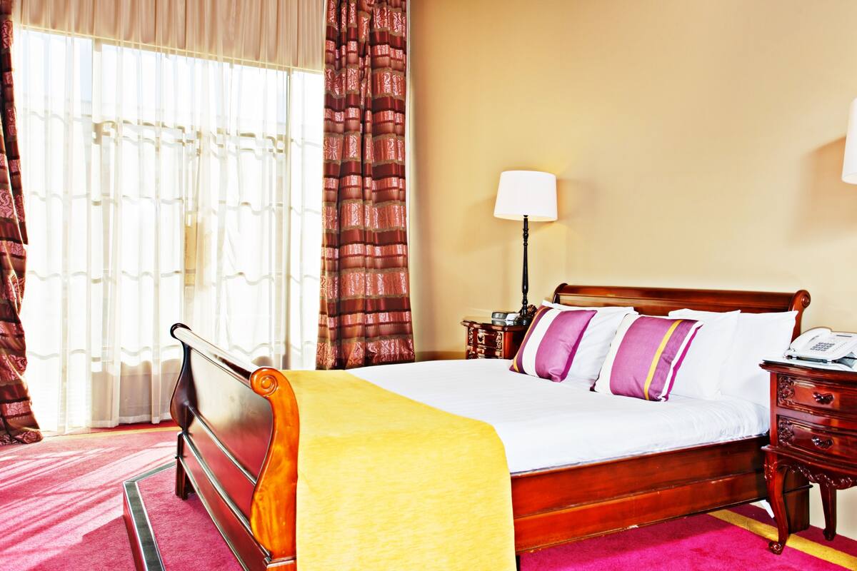 A hotel room with a wooden bed, a bedside table, a lamp, and a window with curtains.