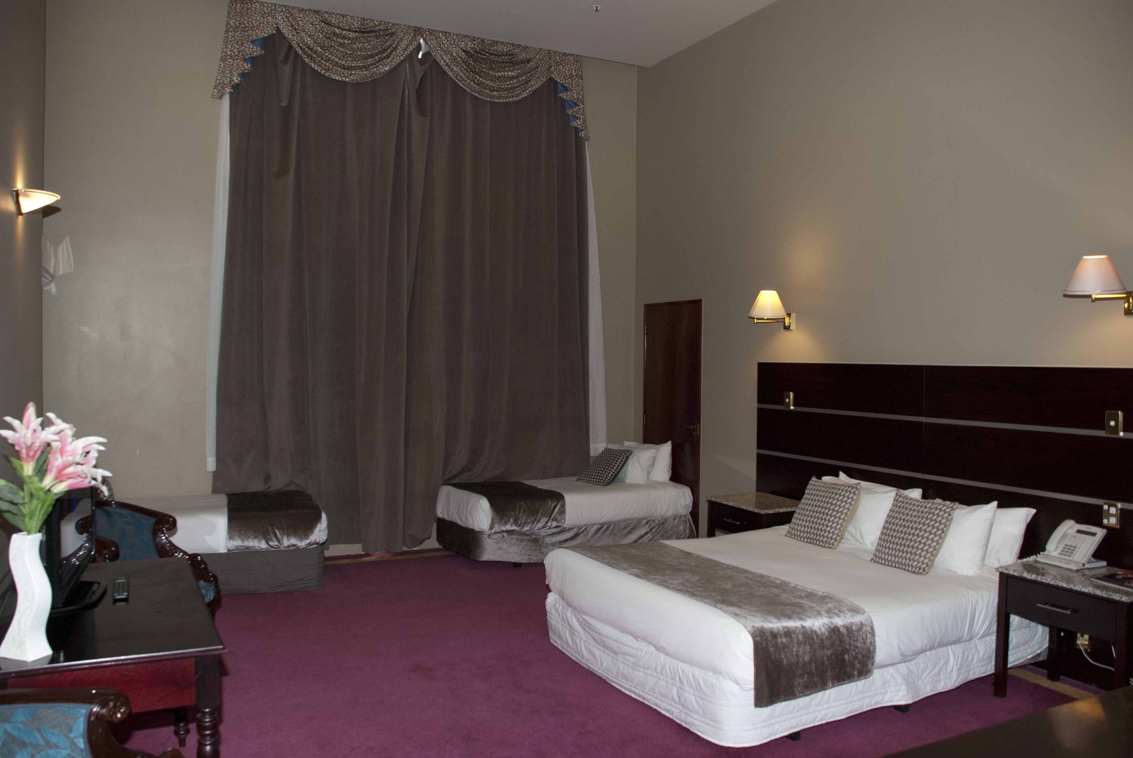 A hotel room with two beds, a desk, a chair, a vase with flowers, and a large window with curtains.
