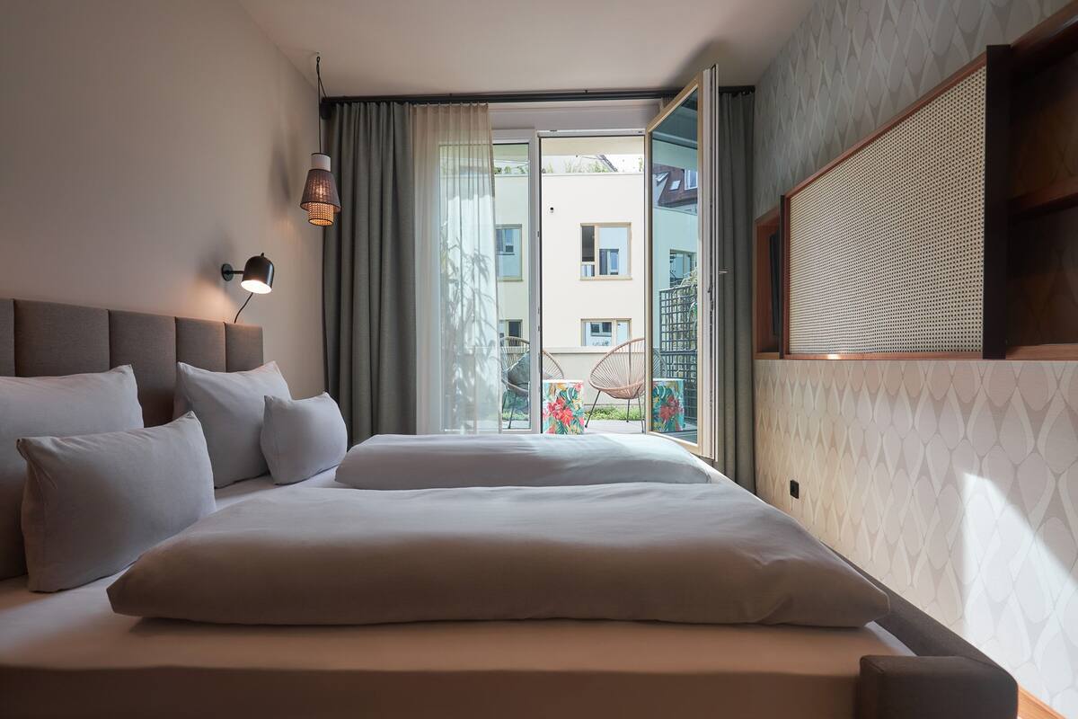 A hotel room with a large bed, a nightstand, a wall-mounted lamp, and a view of the outside balcony.