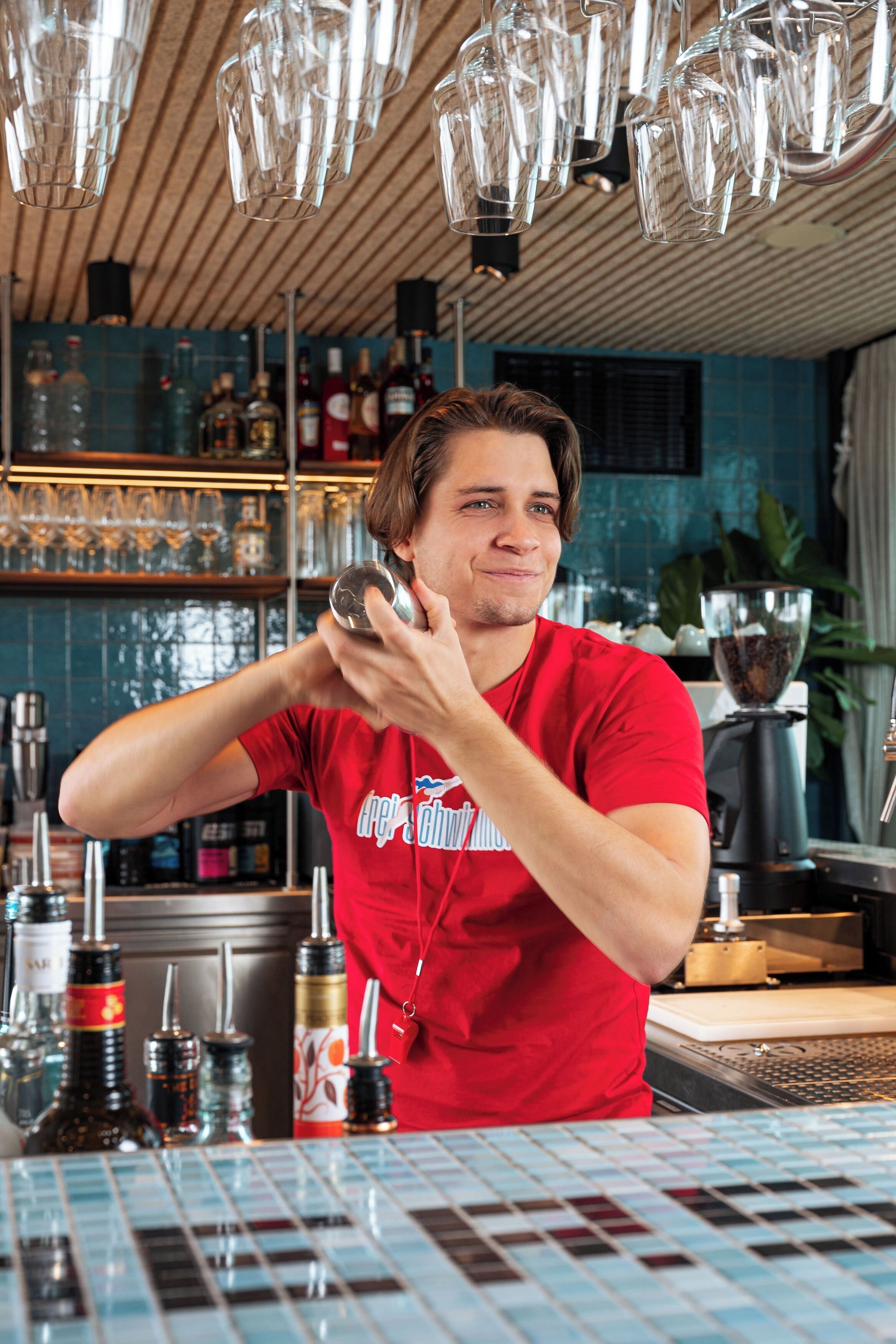 Un barman, vêtu d’une chemise rouge, prépare un cocktail dans un bar dont le mur derrière le comptoir est recouvert de carreaux, et où l’on aperçoit diverses bouteilles en arrière-plan.