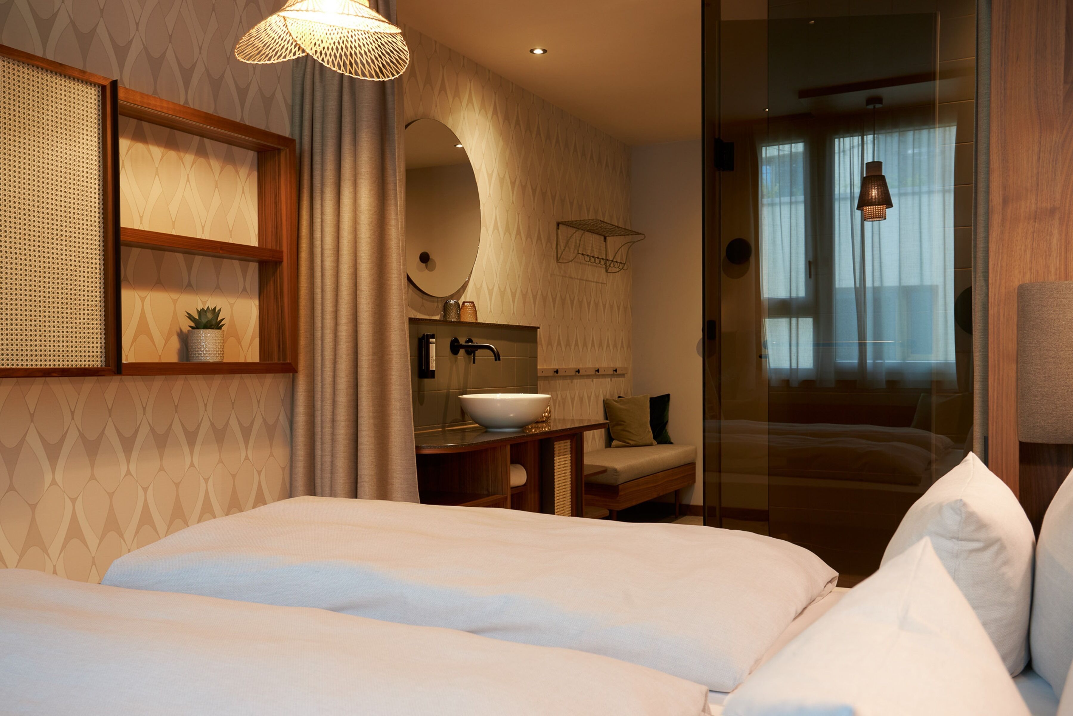 Habitación de hotel con una cama grande, una mesita de noche con lavamanos blanco, un espejo y una planta pequeña. Hay una ventana con cortinas y una lámpara colgante.