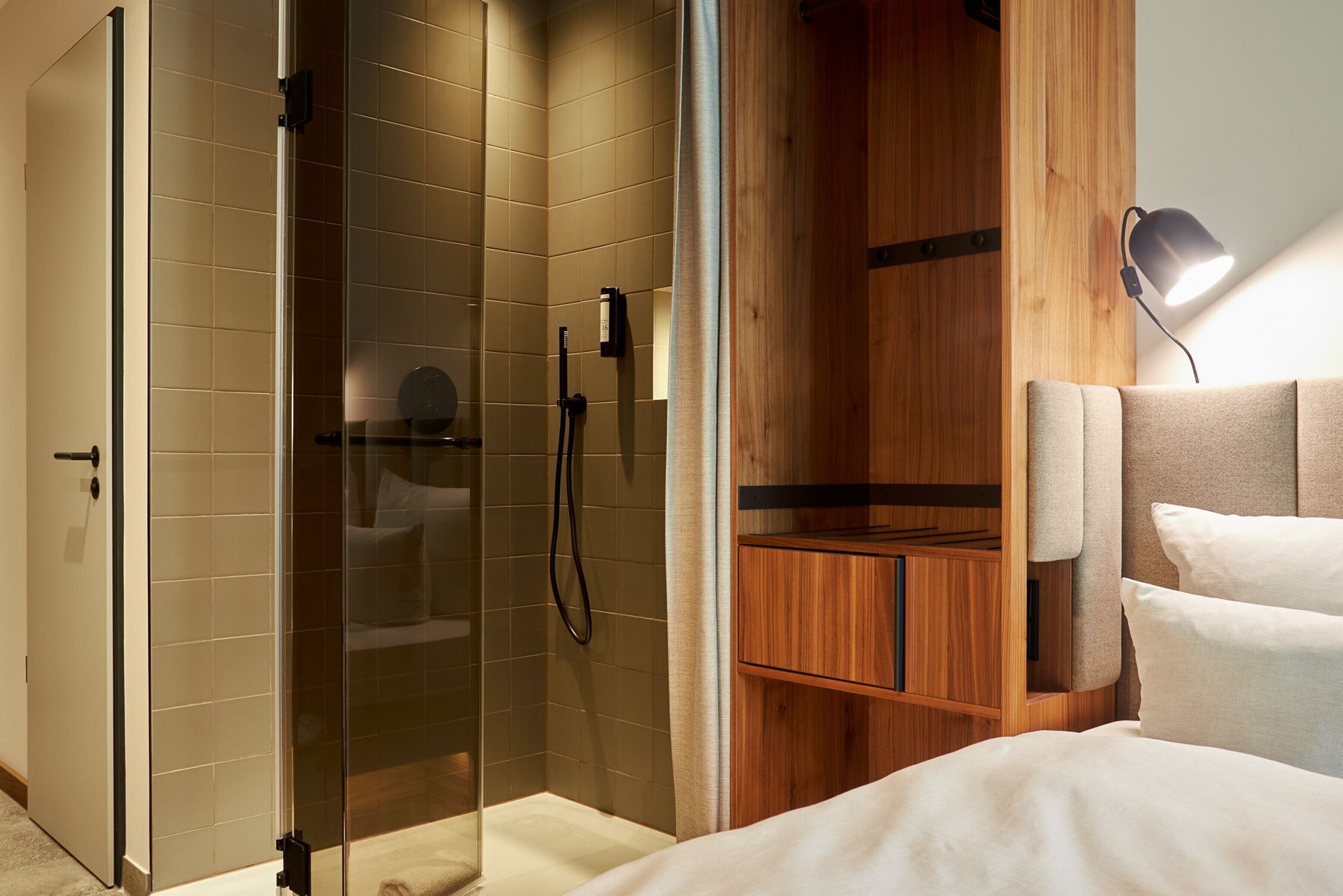 A modern bathroom with a glass shower enclosure, wooden cabinetry, and a bedside lamp.