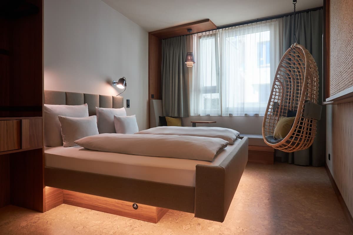 A modern bedroom with a bed, bedside tables, a hanging chair, and a window with curtains.