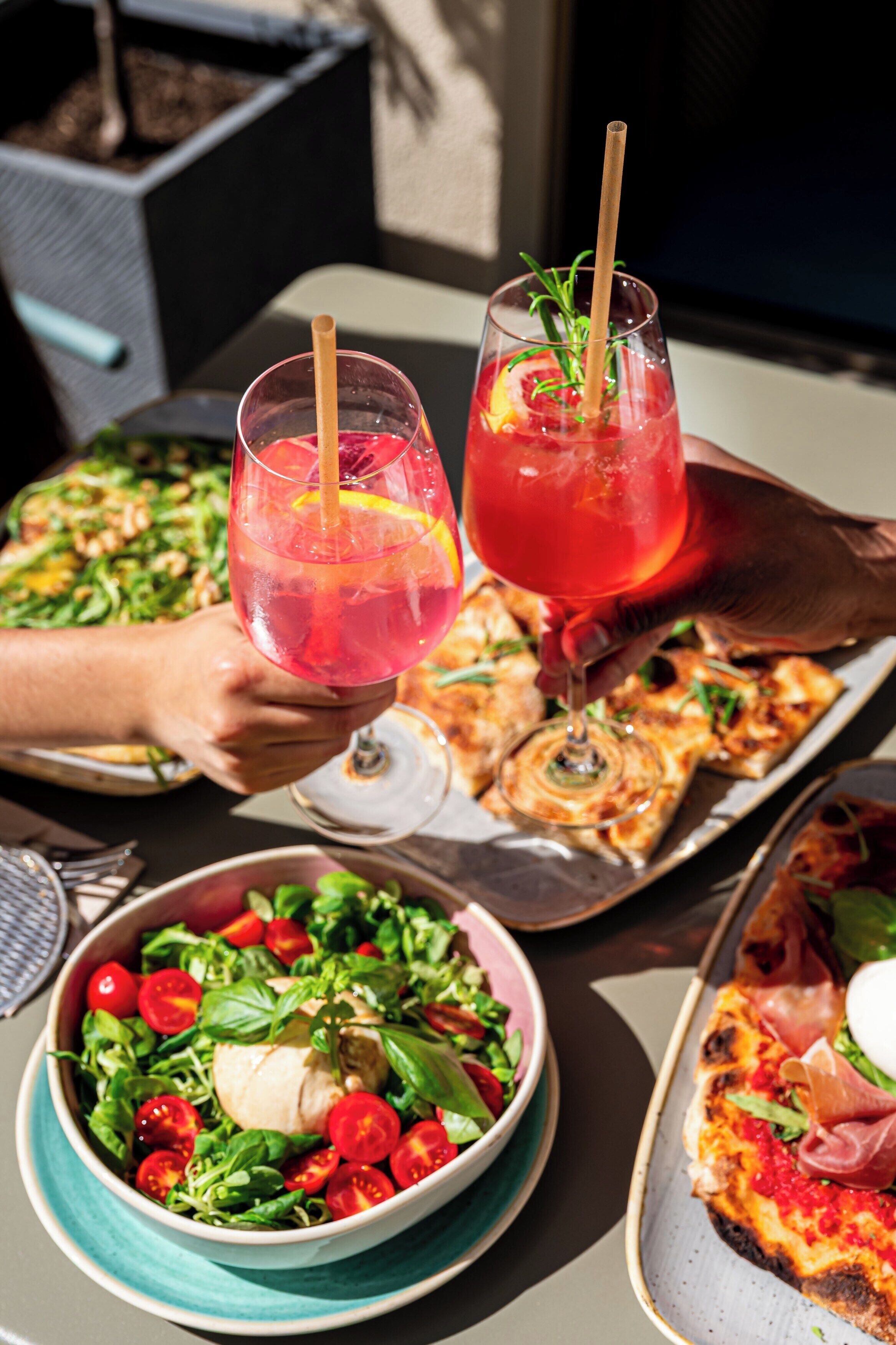 Una mesa con comida que incluye pizza, ensalada y un tazón de vegetales. También se observan dos bebidas con pajitas.