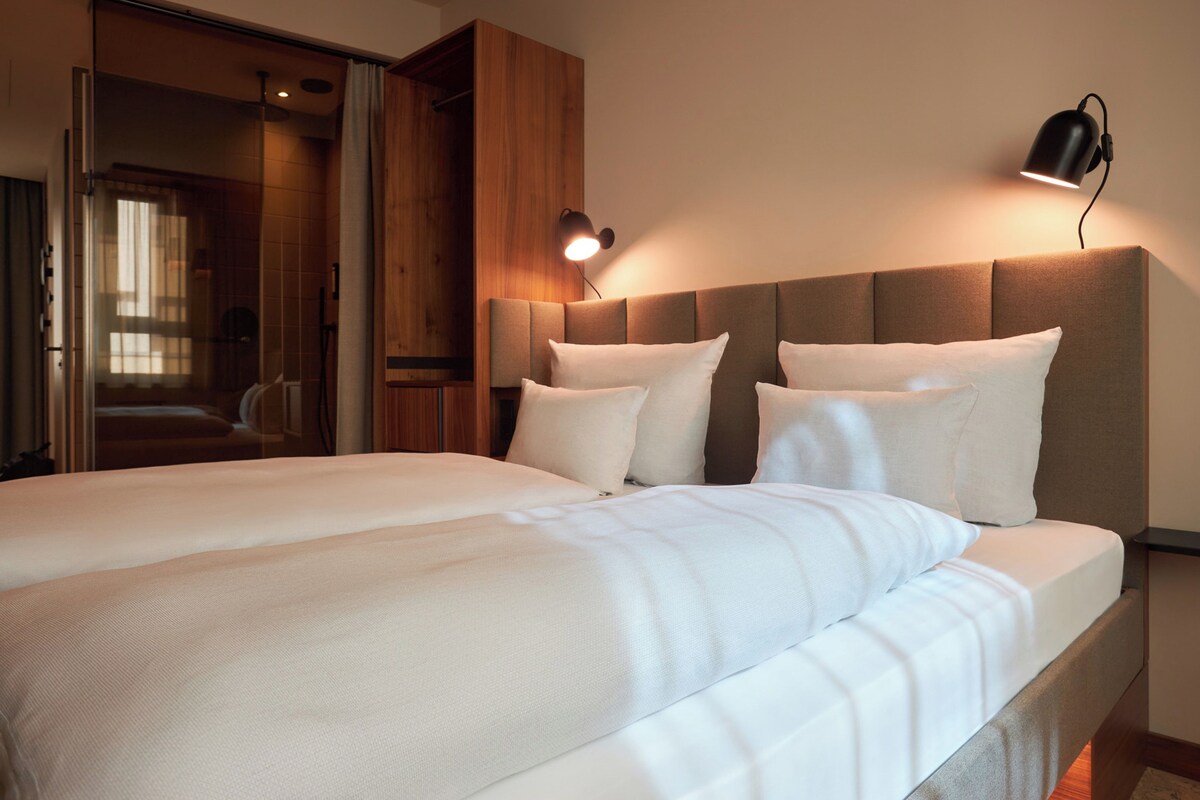 Una habitación de hotel con una cama grande, un armario de madera, un baño con puerta de cristal y una lámpara fijada a la pared.