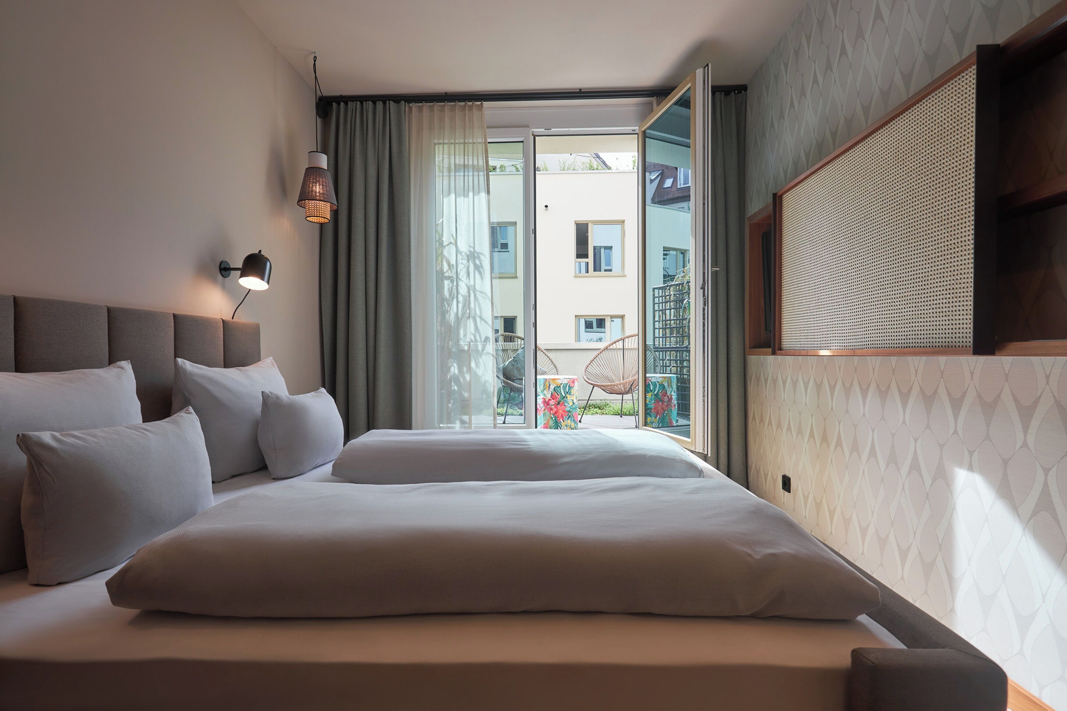 Moderni hotellihuone, jossa on suuri sänky, yöpöytä, seinään kiinnitetty lamppu ja parveke, jolta on näkymä kaupunkiin.