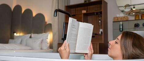 A person reading a book in a bathtub with a modern headboard and a wooden shelf in the background.