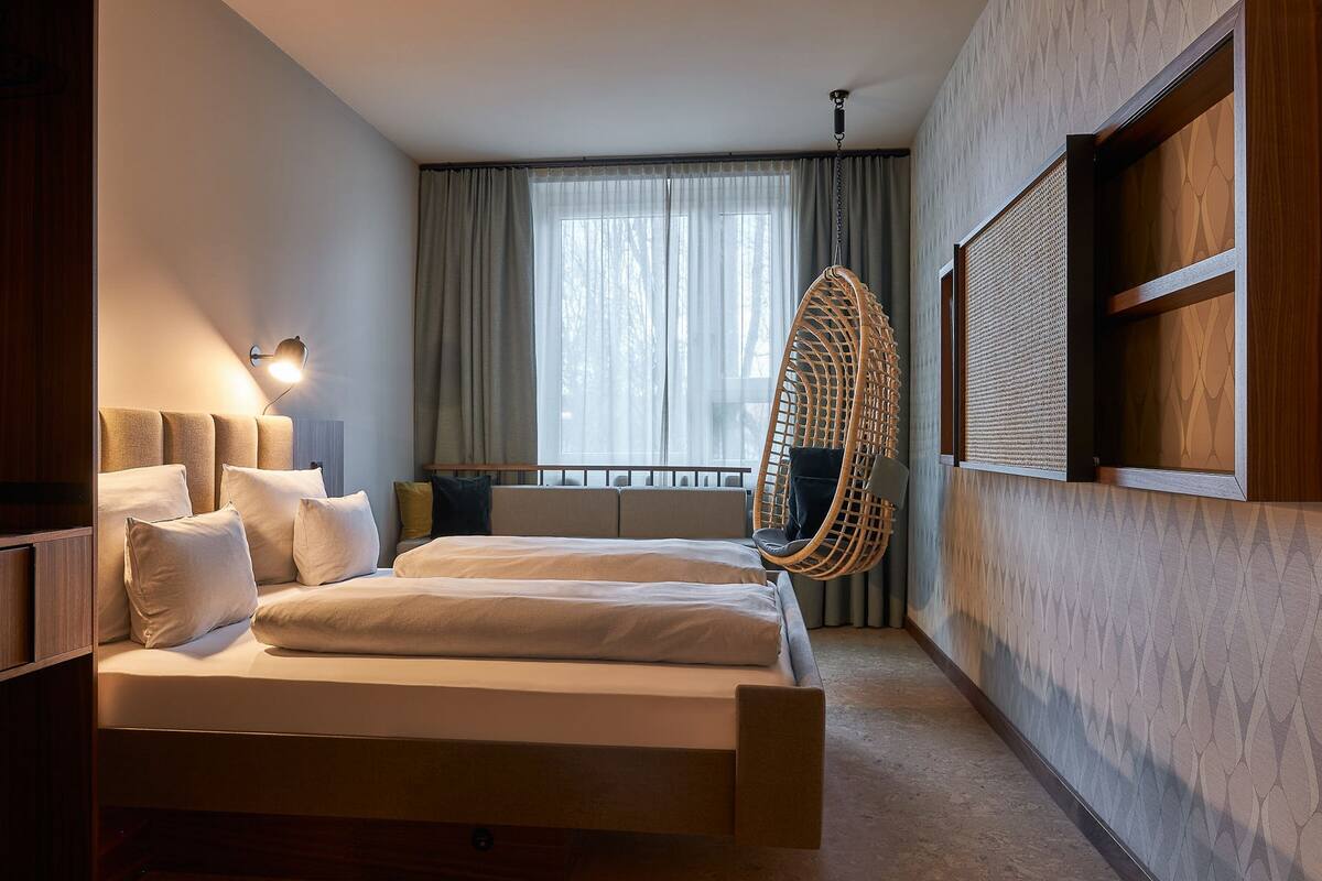 A modern bedroom with a large bed, a hanging chair, a desk, and a window with curtains.