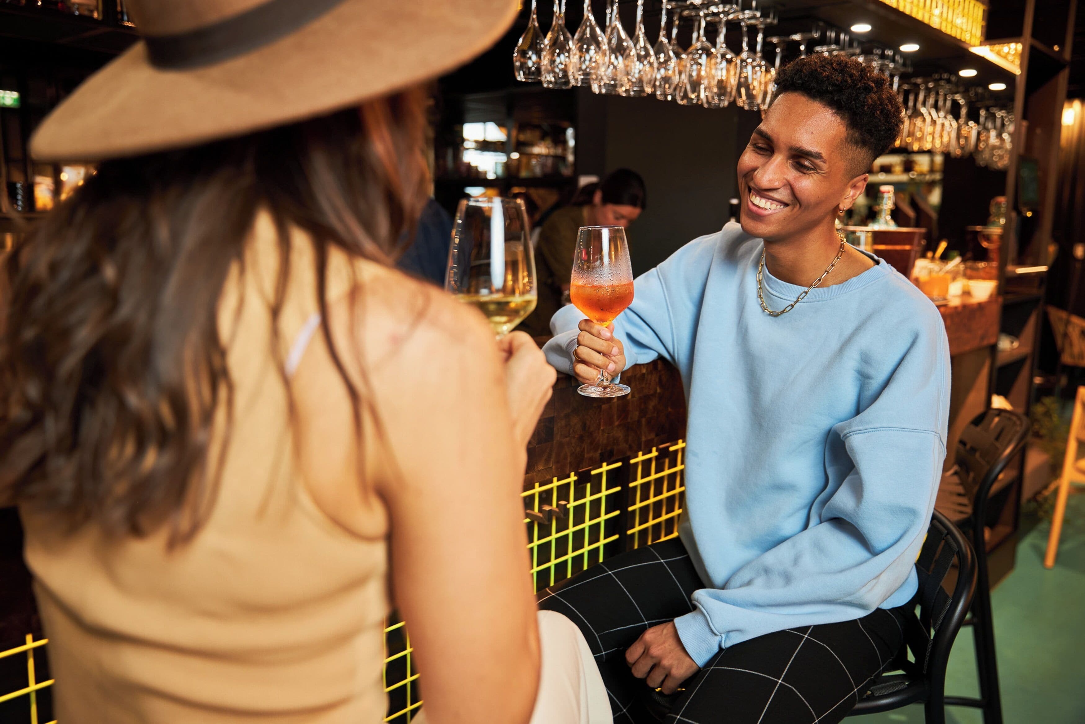 Una persona sosteniendo un vaso de bebida en un bar con copas colgantes y una pared con diseño de cuadrícula al fondo.