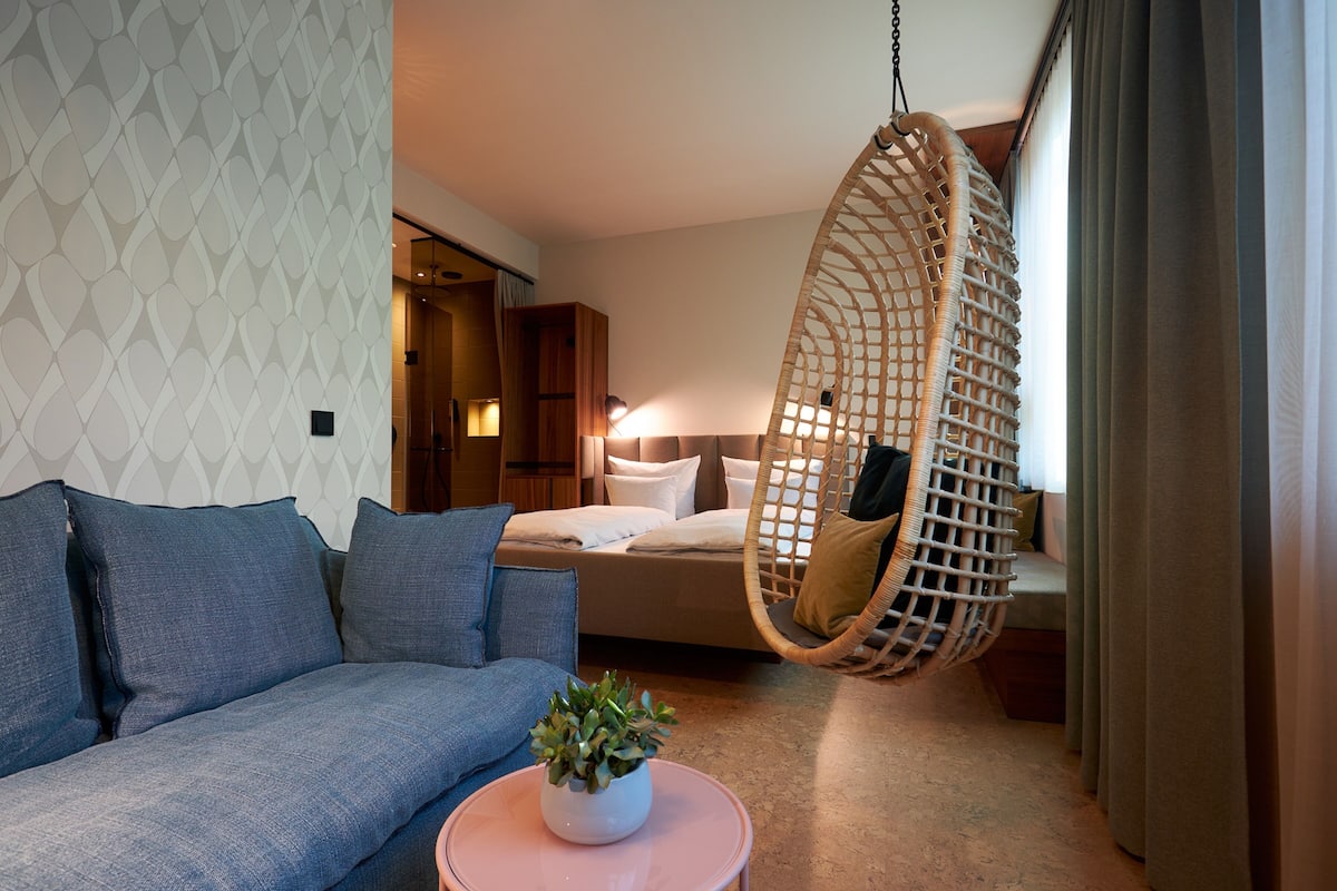 A modern hotel room with a hanging chair, a sofa, a bed, and a small table with a plant.