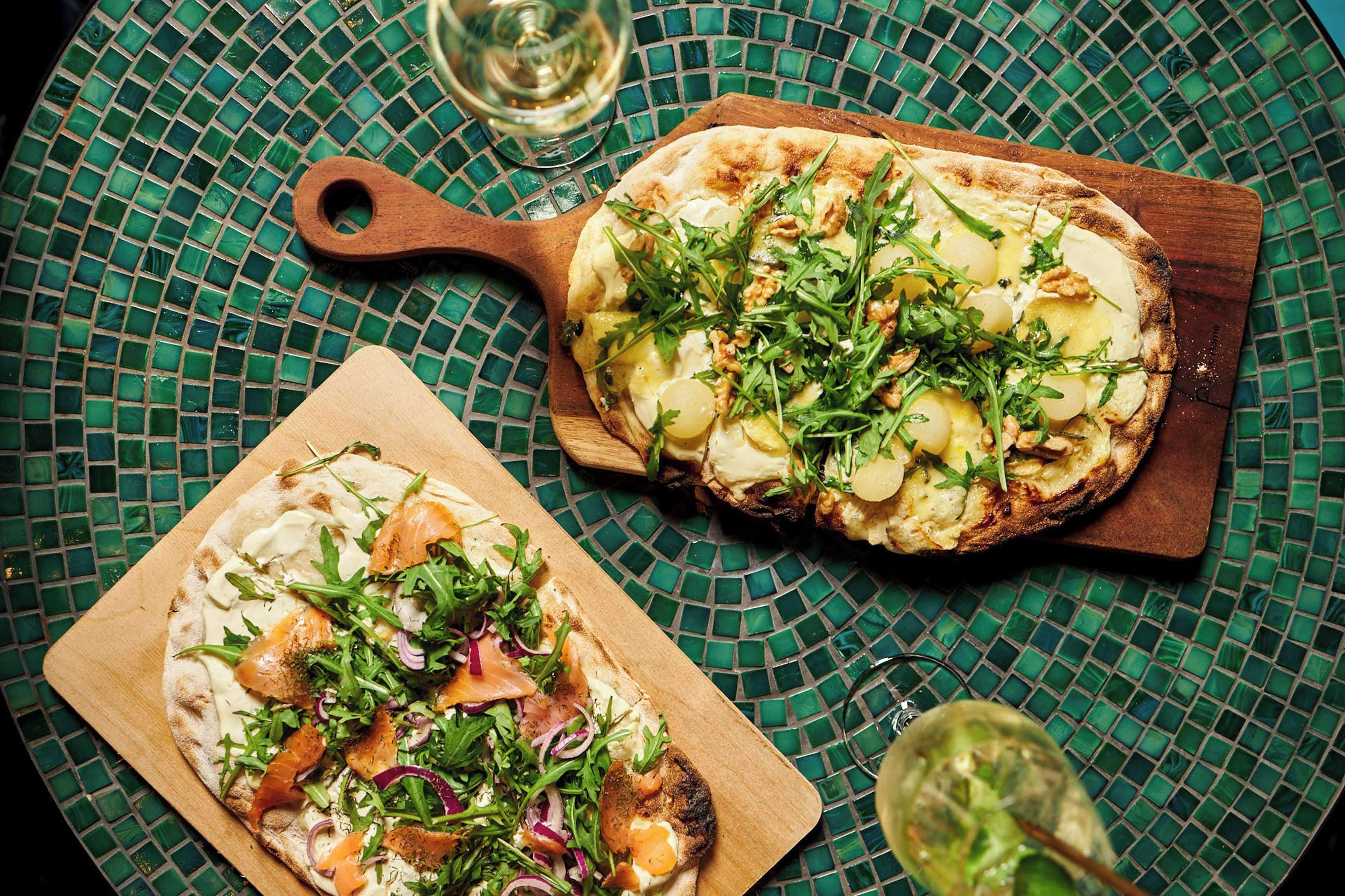 Two pizzas on wooden boards with a green mosaic tablecloth.