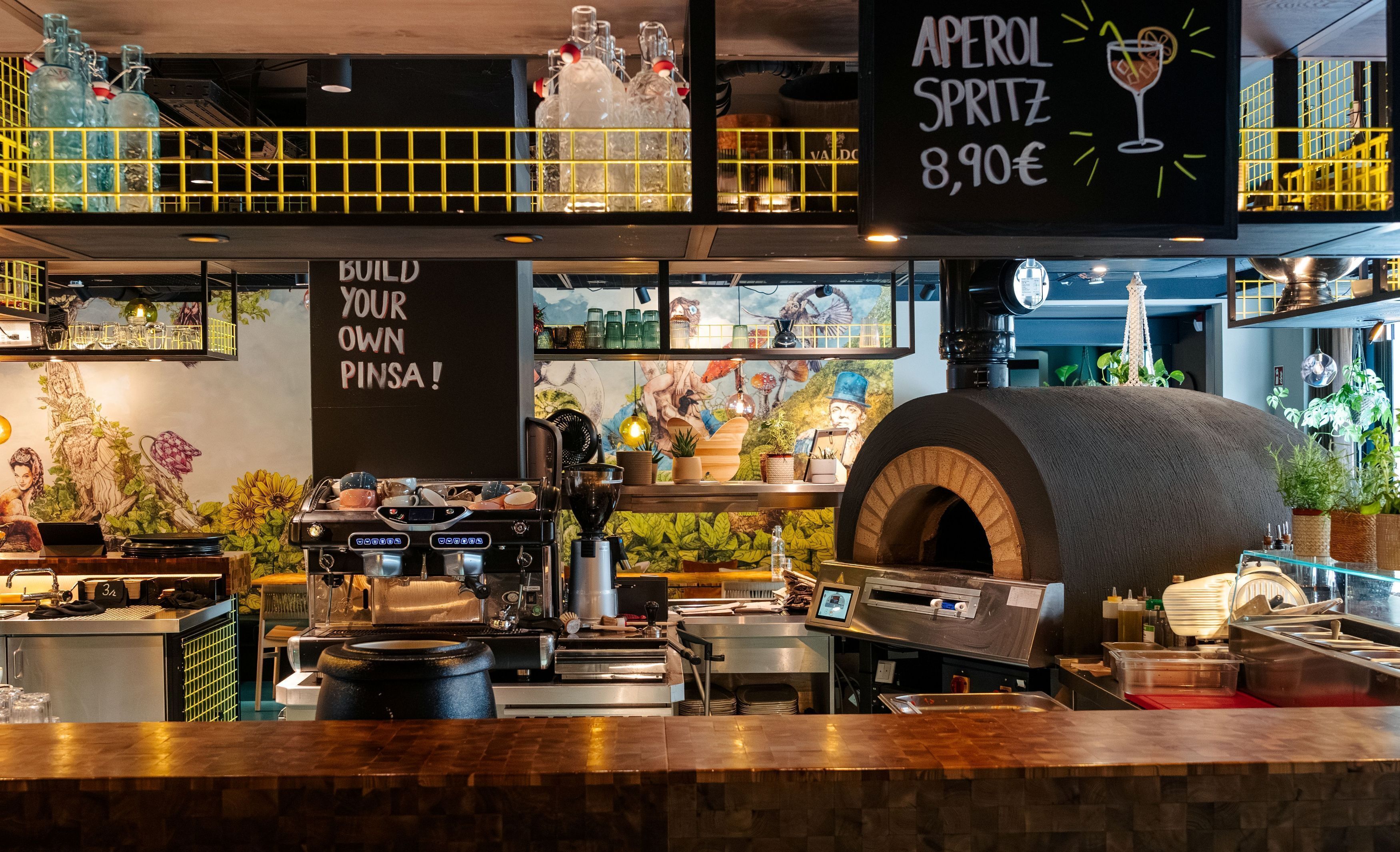 A coffee shop interior with espresso machines, a large oven, and a menu board advertising 'APEROL SPRITZ' for 8,90€.
