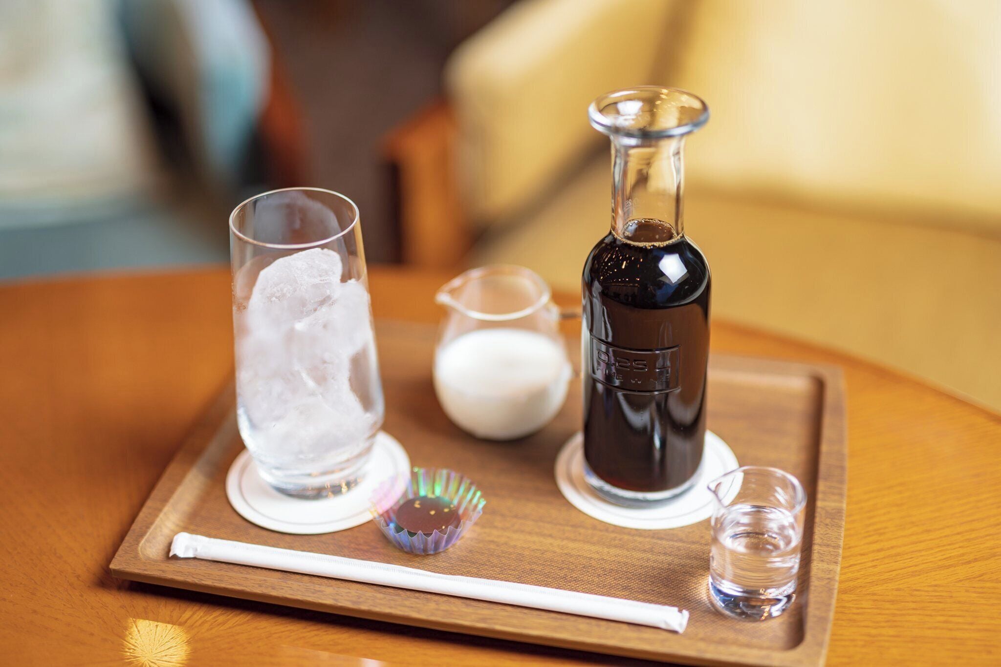 A tray with a glass of iced water, a bottle of dark liquid, a small pitcher, and a shot glass on a wooden table.