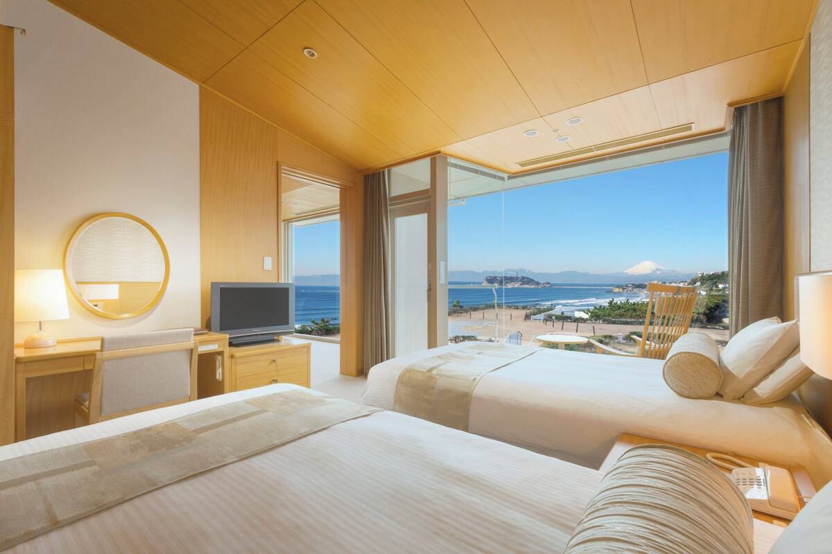 A hotel room with two beds, a TV, a desk, and a large window with a view of the beach and mountains.
