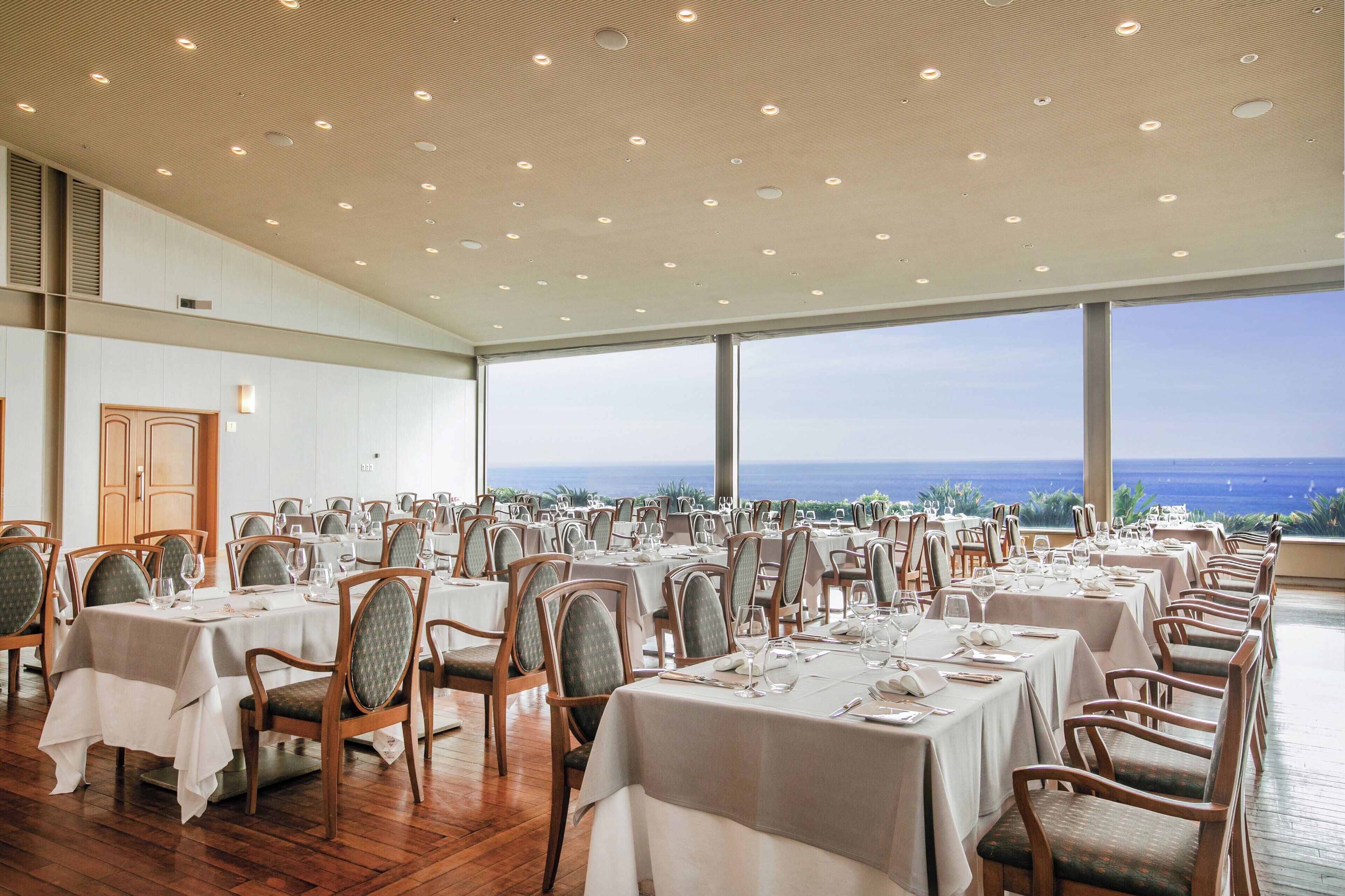 A spacious dining area with multiple tables and chairs, large windows offering a view of the ocean, and a high ceiling with recessed lighting.