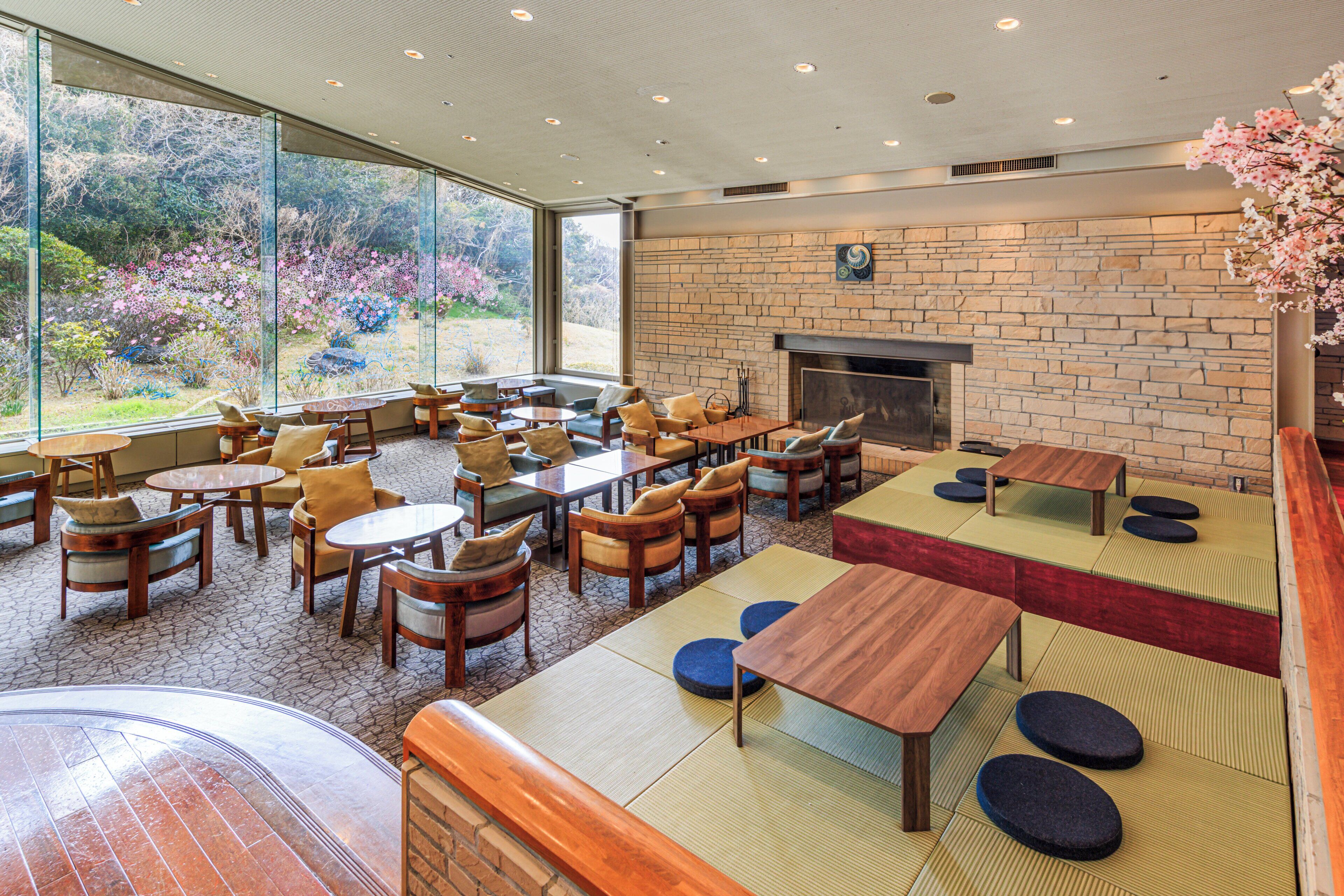 A spacious indoor area with multiple seating arrangements, including tables and chairs, a fireplace, and large windows offering a view of a garden.
