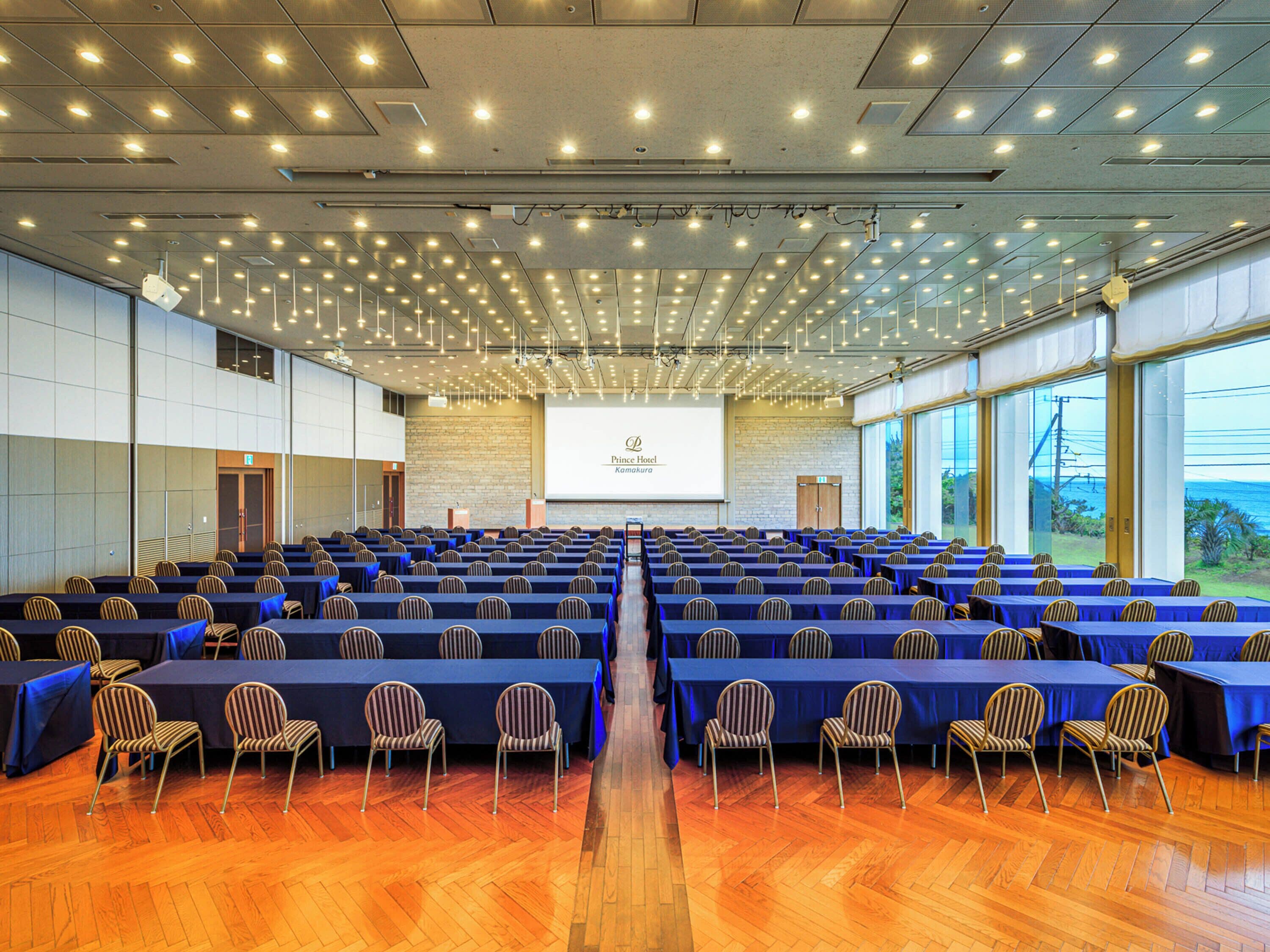 A conference room with rows of tables and chairs, a large screen, and a view of the ocean.