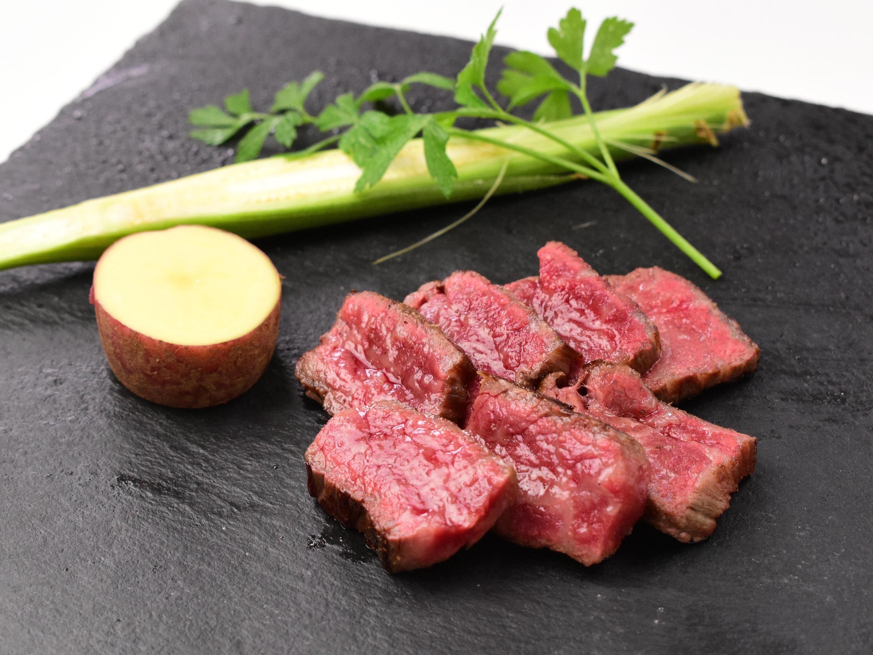 Une planche de service en ardoise noire, sur laquelle on a disposé des tranches de bœuf, une pomme de terre coupée en deux et un bouquet d’herbes fraîches.