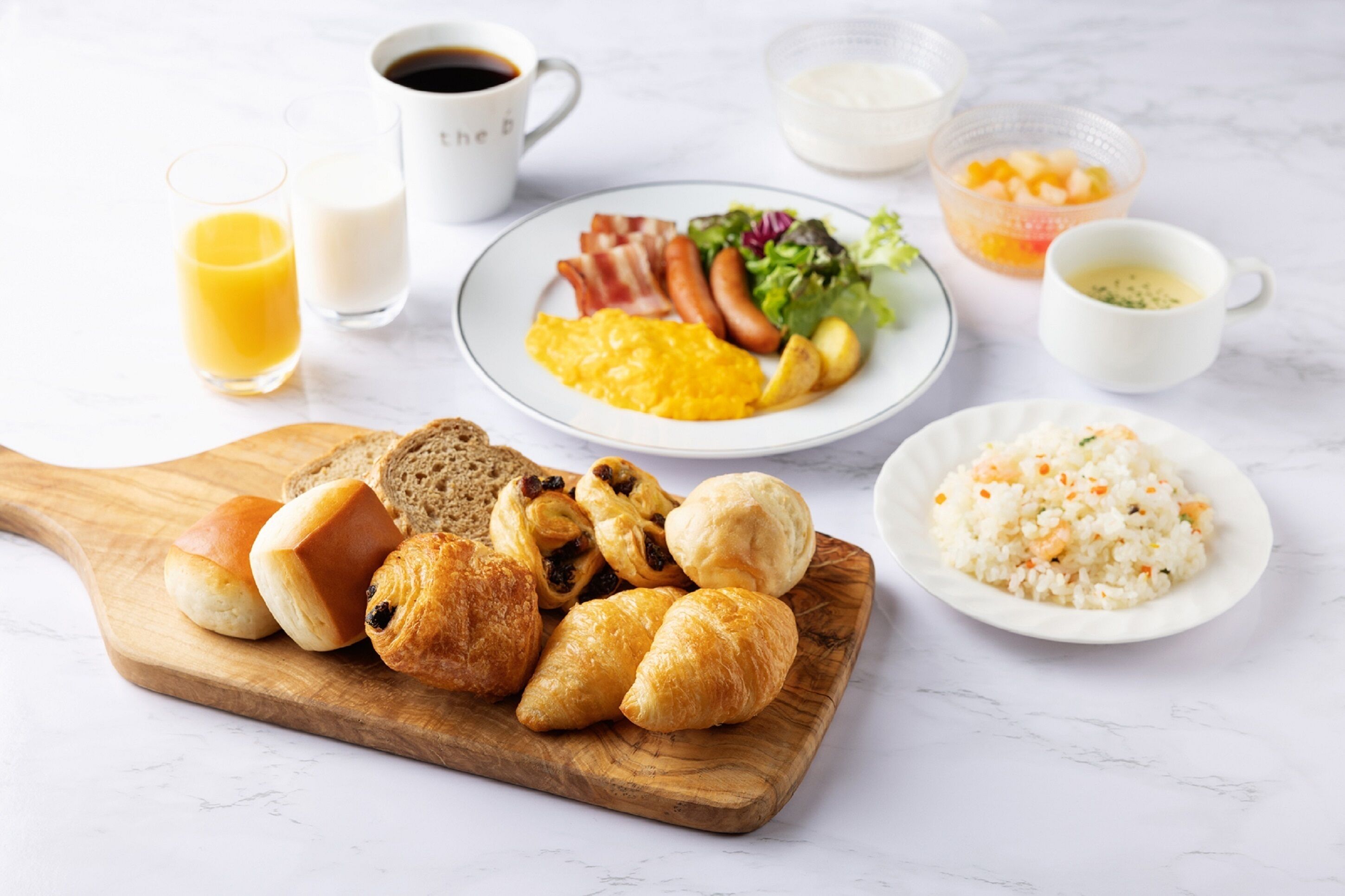 A breakfast table with croissants, bread, scrambled eggs, bacon, sausages, a salad, and rice. There are also two glasses of orange juice and a cup of coffee.