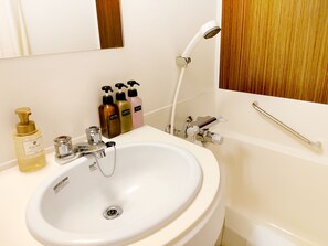 A bathroom with a white sink, a mirror, a handheld showerhead, and three bottles of shampoo and conditioner. - the b hachioji (Hachioji)