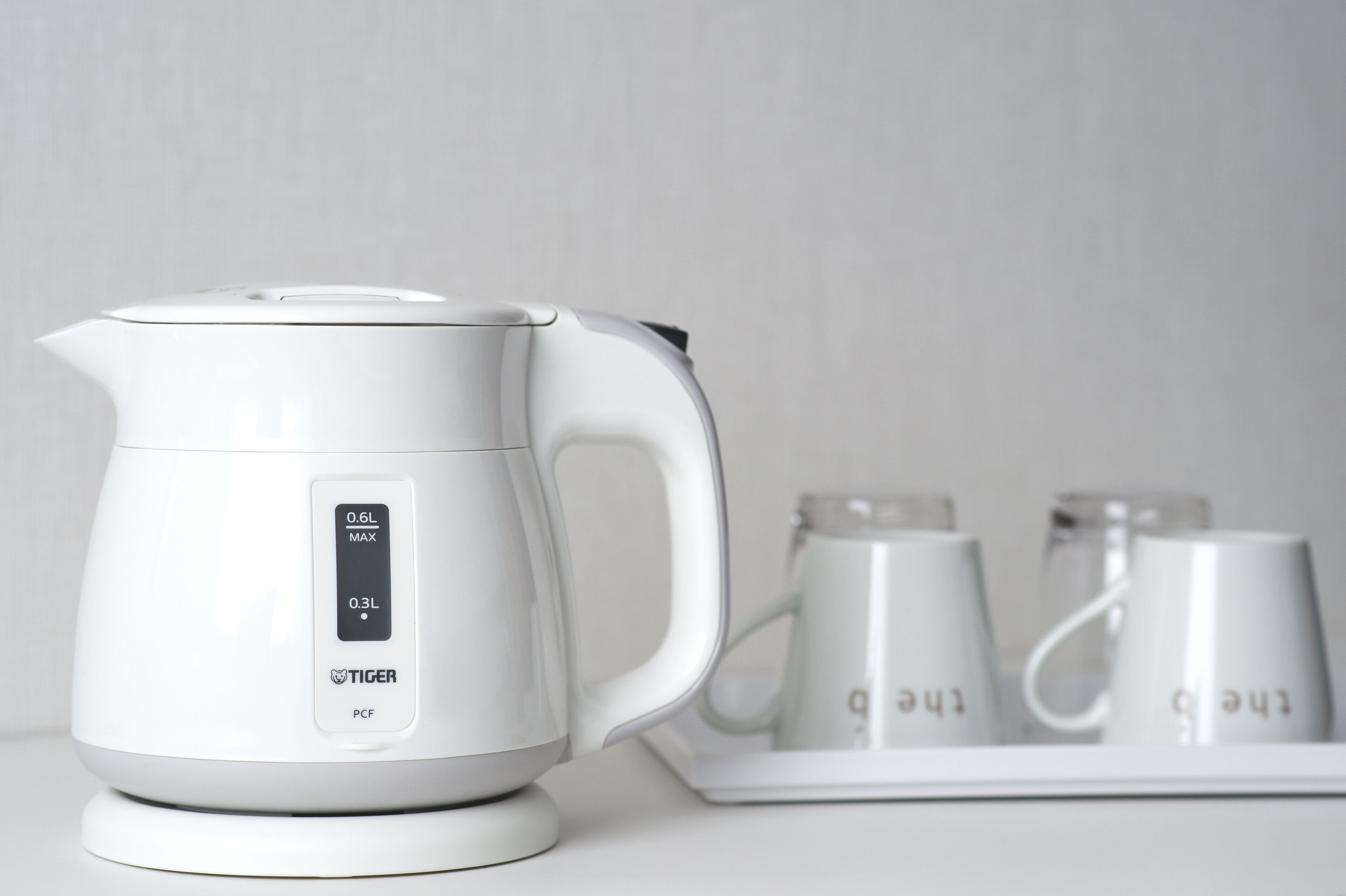 A white electric kettle with a digital display, placed on a white surface with two white mugs in the background.