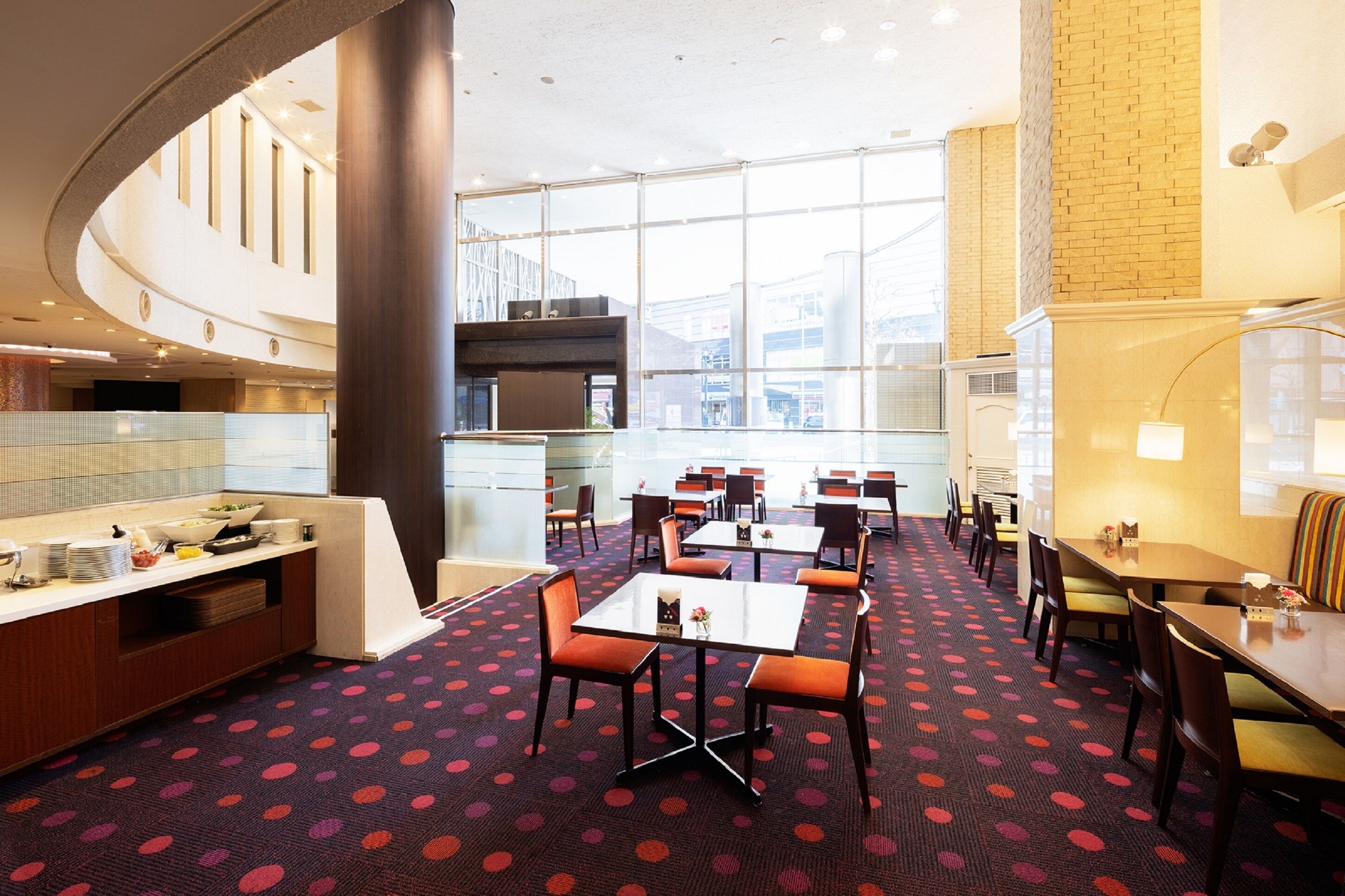 En hotellobby med modern design, med en central sittgrupp med bord och stolar, ett köksområde och stora fönster som erbjuder utsikt över staden.