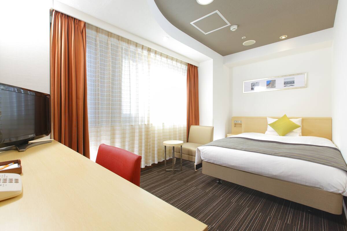 Habitación de hotel con cama, escritorio, silla, televisor y ventana con cortinas.