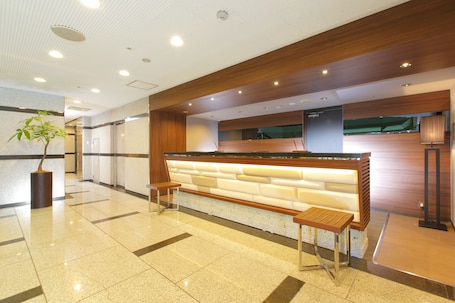A reception area with a wooden reception desk, two wooden stools, a potted plant, and a tiled floor.