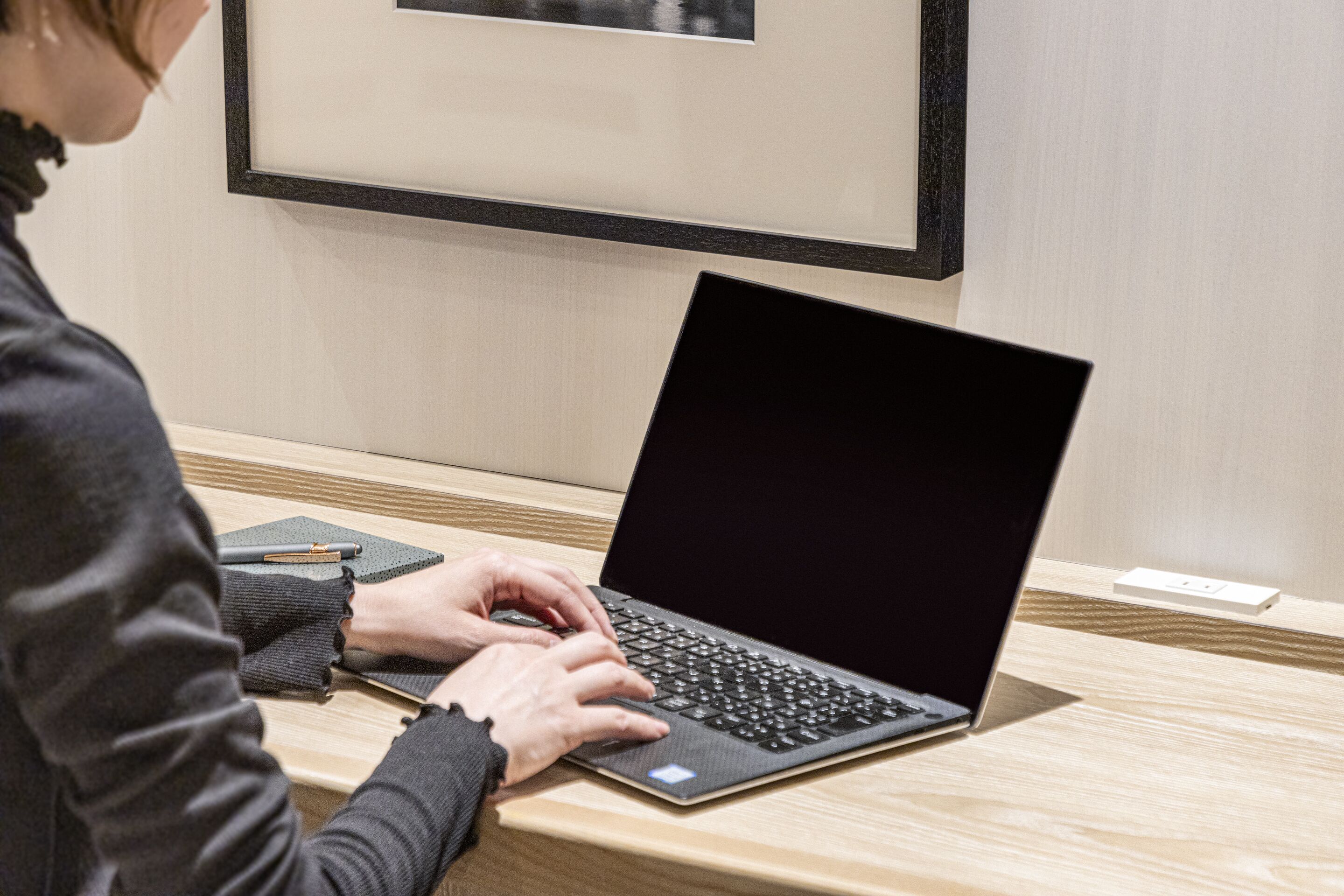 Une personne utilise un ordinateur portable sur un bureau en bois, et un tableau encadré est accroché au mur.