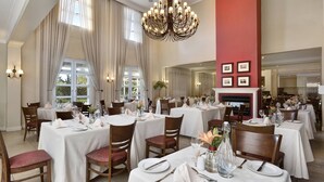 A dining area with tables set for service, large windows with curtains, and a chandelier.